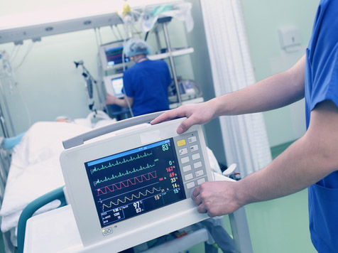 Picture of a heart monitor machine in pre-op at the foot of the bed. There is a female Nurse in the background putting patient info into a computer and patient lying down next to the Nurse.