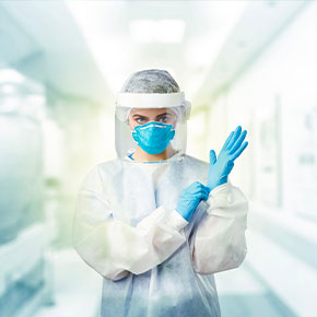Picture of a female Nurse wearing a surgical cover, head cover, face shield, mask, and gloves.She is standing in a bright hall way/