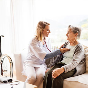 Picture of a female in home Nurse sitting on a couch next to an elderly female patient. She is using her stethoscope to check the elderly woman&apos;s heart beat. They are both smiling at each other.