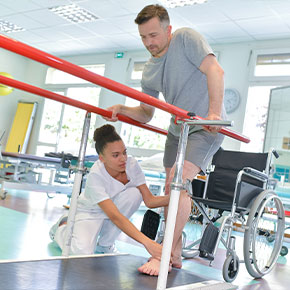 Picture of a female Nurse helping assist a male patient. He is standing up from his wheel chair and grabbing hand rails for support to try and start walking. The female Physical Therapist is pushing his foot down to the ground and holding his knee straight.